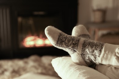 Photo of Woman in knitted socks resting near fireplace at home, closeup of legs