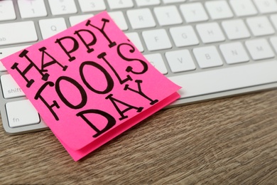 Photo of Note with phrase Happy Fools' Day and keyboard on wooden table, closeup