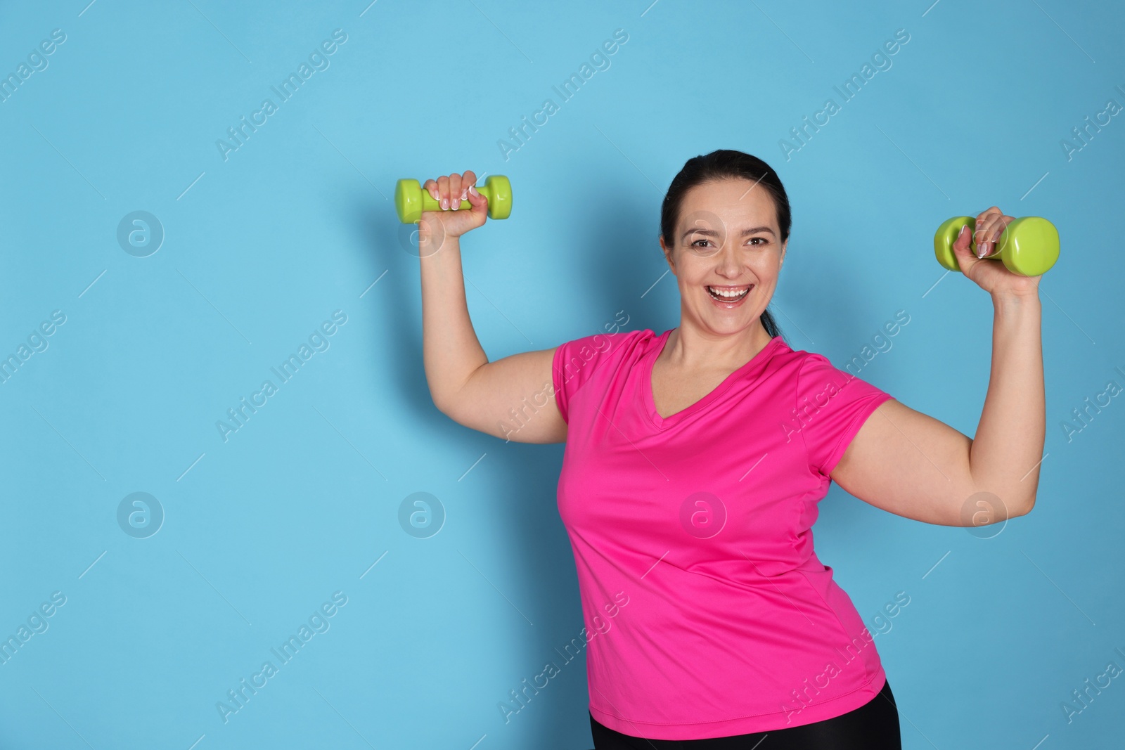 Photo of Happy overweight woman doing exercise with dumbbells on light blue background, space for text