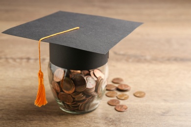 Graduation hat and jar with coins on wooden table, space for text. Money saving concept