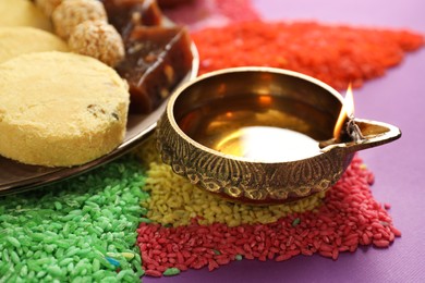 Photo of Diwali celebration. Tasty Indian sweets, diya lamp and colorful rangoli on violet table, closeup