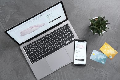 Photo of Online store website on laptop screen. Computer, smartphone, credit cards and houseplant on grey table, flat lay
