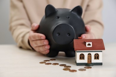 Man holding piggy bank near house model and coins at wooden table, closeup. Saving money concept