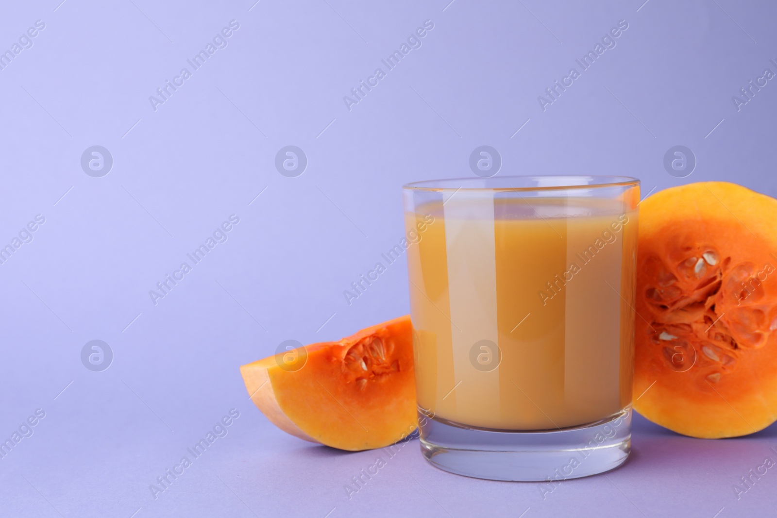 Photo of Tasty pumpkin juice in glass and cut pumpkin on lavender color background. Space for text