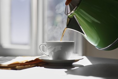 Photo of Pouring hot drink into cup near newspaper on sunlit table. Good morning