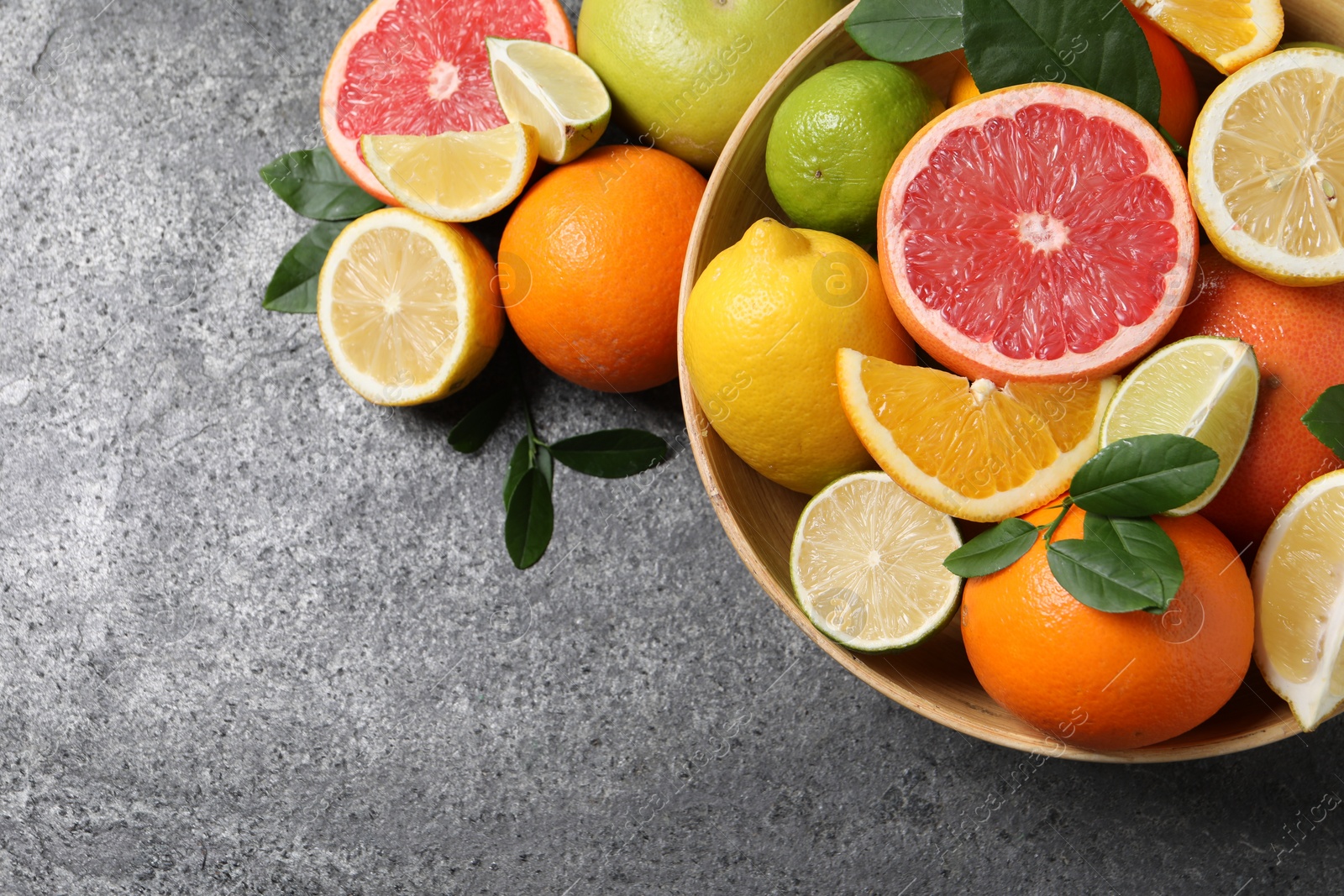 Photo of Different fresh citrus fruits and leaves in bowl on grey textured table, flat lay. Space for text