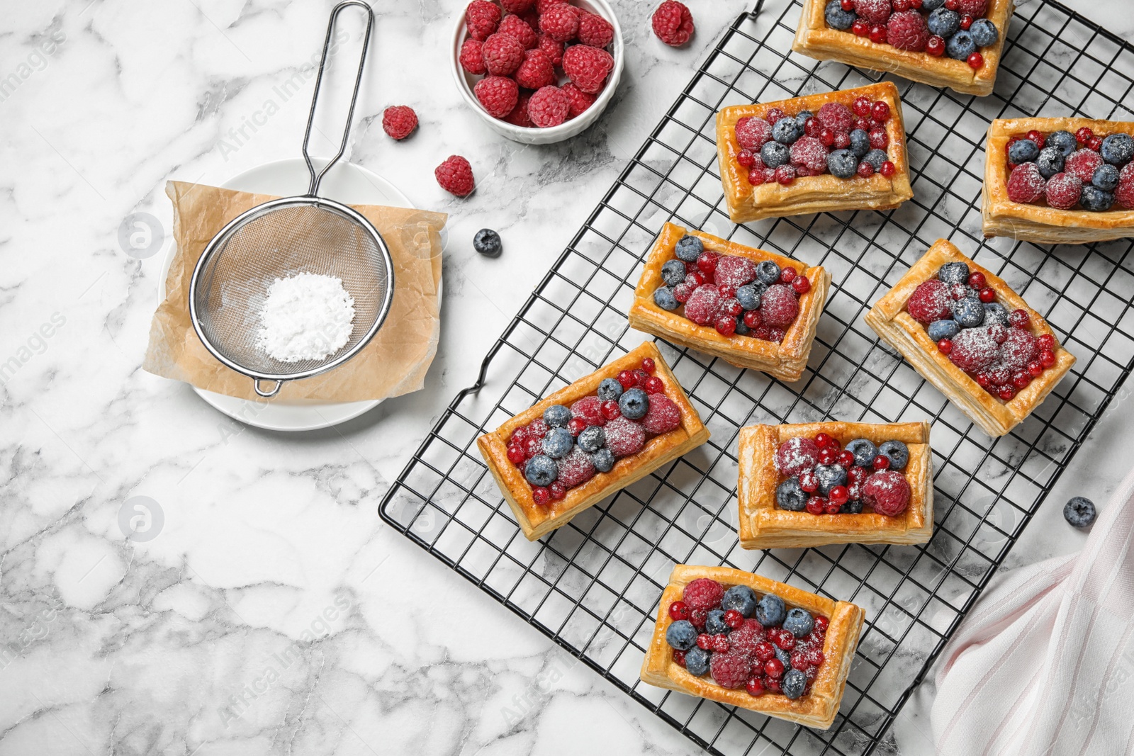 Photo of Cooling rack and fresh delicious puff pastry with sweet berries on white marble table, flat lay