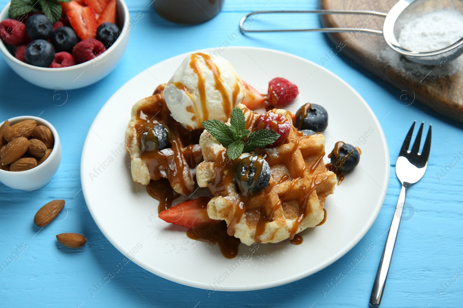 Photo of Delicious Belgian waffles with ice cream, berries and caramel sauce on light blue wooden table, closeup