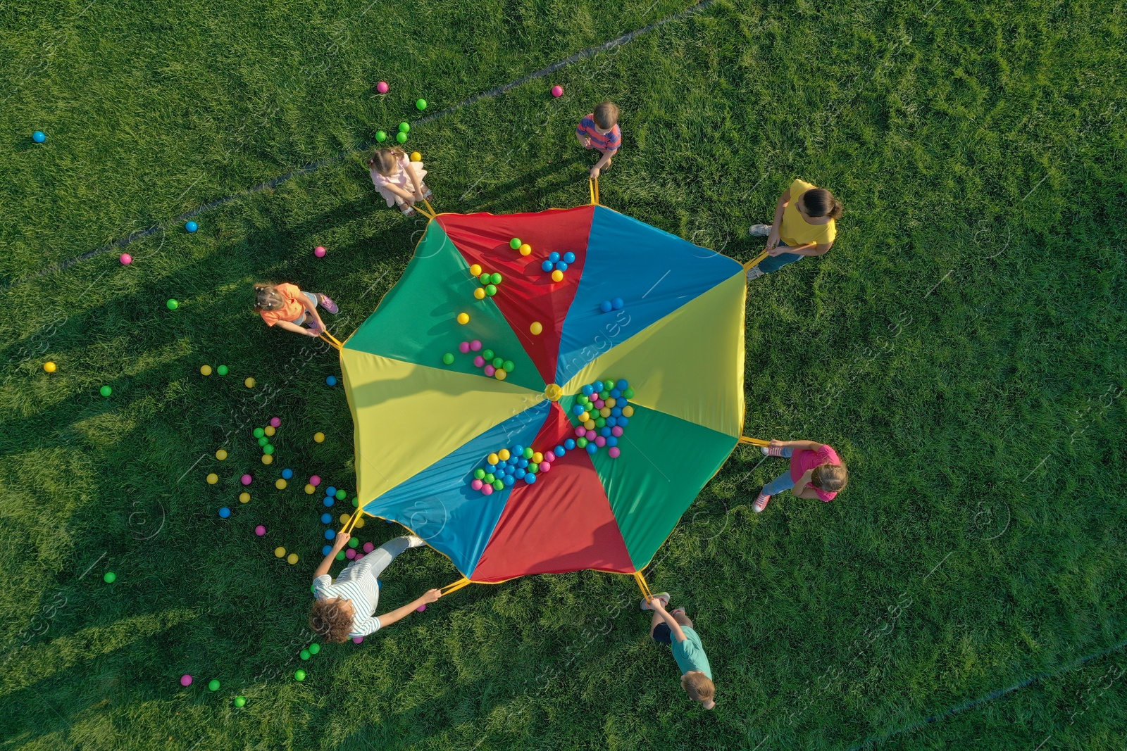 Photo of Group of children and teachers playing with rainbow playground parachute on green grass, top view. Summer camp activity