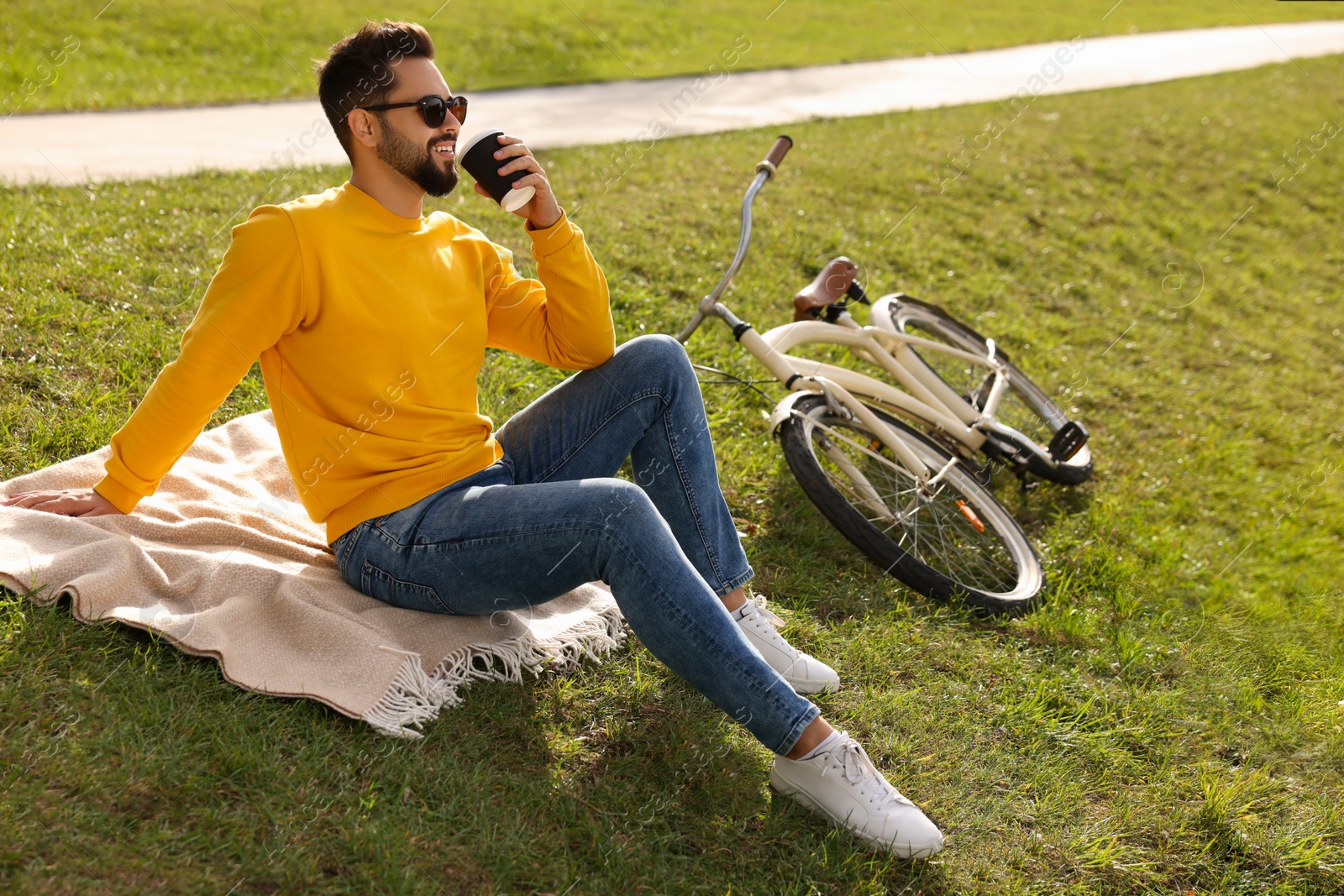 Photo of Young man sitting on green grass and drinking coffee near bicycle outdoors, space for text