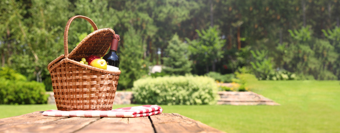 Picnic basket with fruits, bottle of wine and checkered blanket on wooden table in garden, space for text. Banner design