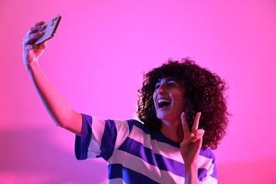 Beautiful young woman taking selfie on color background in neon lights