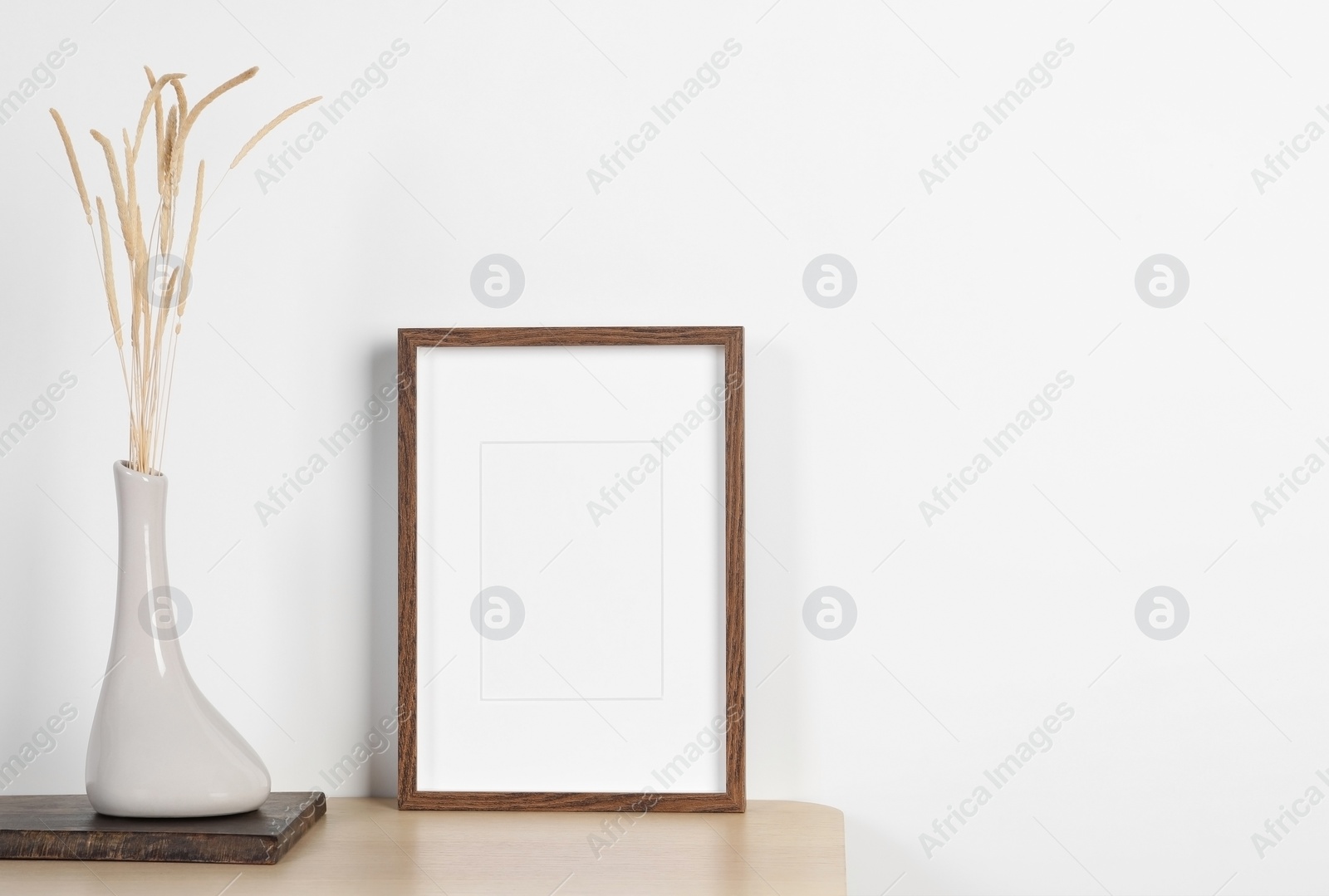 Photo of Empty photo frame and vase with dry decorative spikes on wooden table. Mockup for design