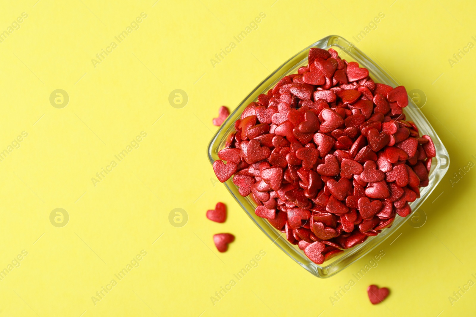 Photo of Bright heart shaped sprinkles in glass bowl on yellow background, flat lay. Space for text