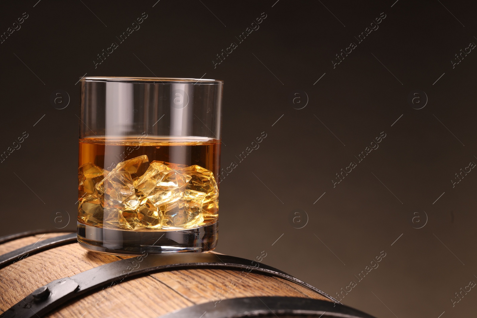 Photo of Whiskey with ice cubes in glass on wooden barrel against dark background, closeup. Space for text