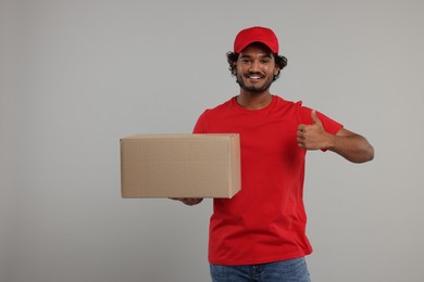 Photo of Happy young courier with parcel showing thumb up on grey background, space for text