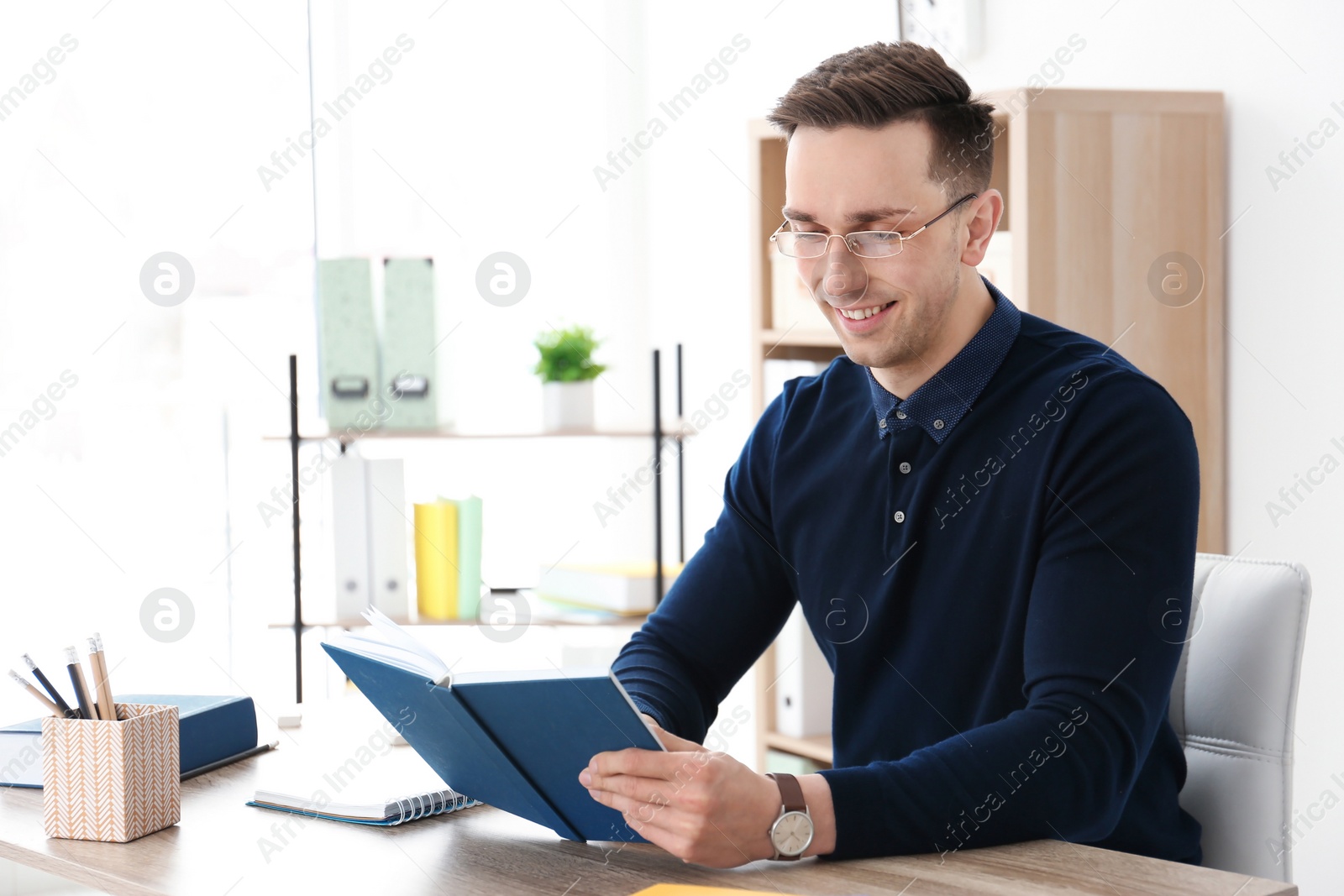 Photo of Young male teacher with book sitting at table in classroom