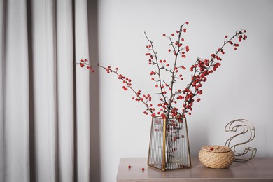 Photo of Hawthorn branches with red berries in vase, decorative letter and wicker box on wooden table indoors, space for text