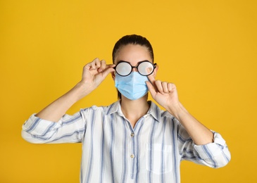Woman wiping foggy glasses caused by wearing medical mask on yellow background