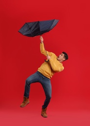 Emotional man with umbrella caught in gust of wind on red background