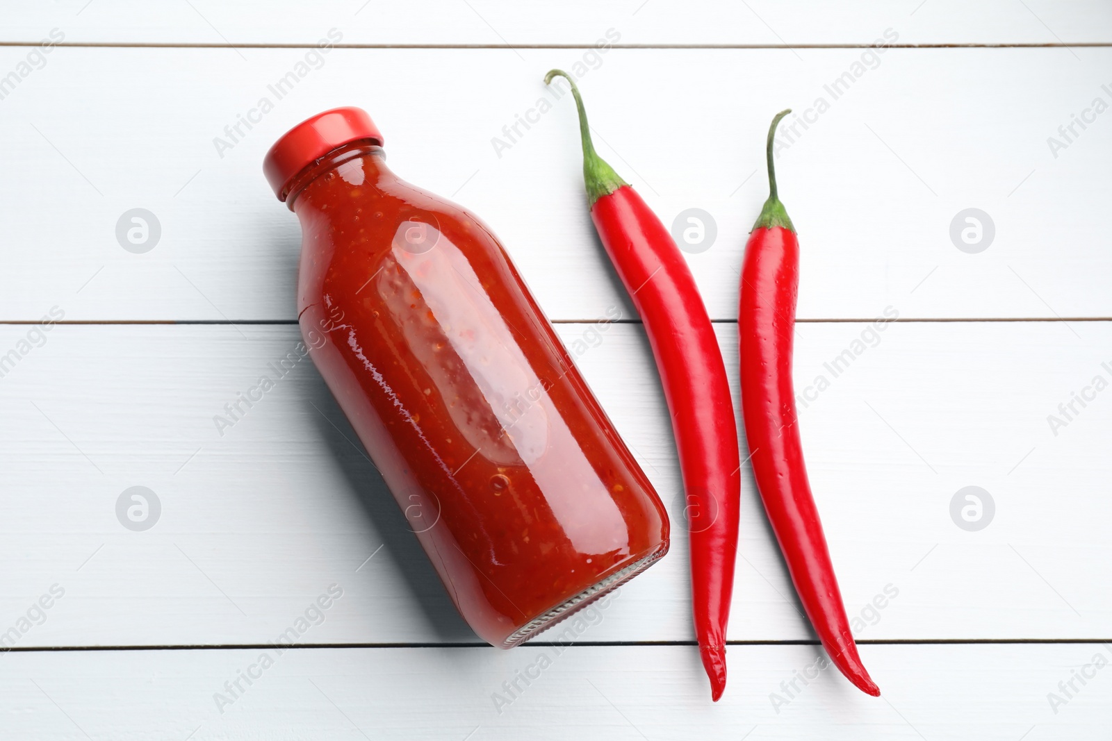 Photo of Spicy chili sauce in bottle and peppers on white wooden table, flat lay