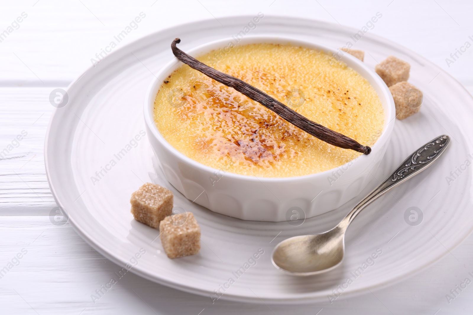 Photo of Delicious creme brulee in bowl, vanilla pod, sugar cubes and spoon on white wooden table, closeup