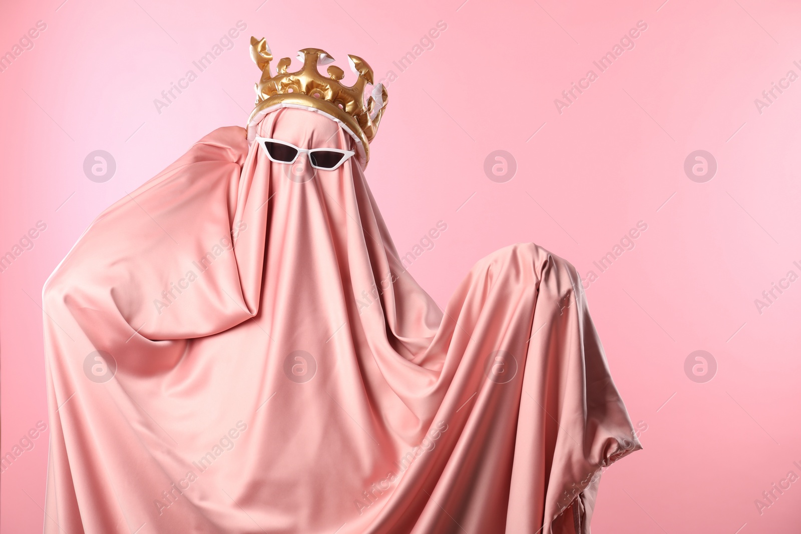 Photo of Glamorous ghost. Woman in sheet with sunglasses and crown on pink background, space for text