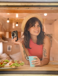 Photo of Young woman with cup and vintage camera in trailer, view from outside. Camping vacation