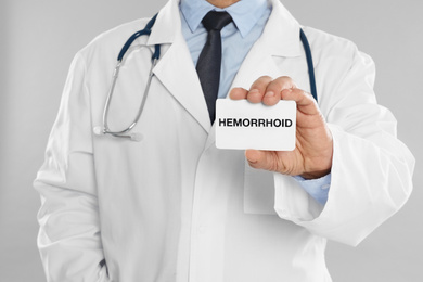 Doctor holding business card with word HEMORRHOID on light grey background, closeup