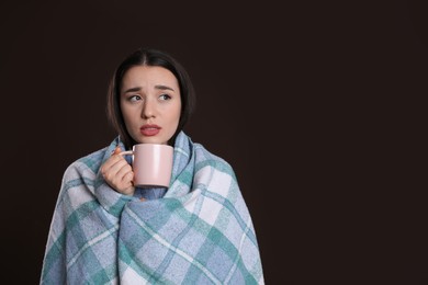 Young woman wrapped in blanket with cup of hot drink suffering from cold on dark background, space for text