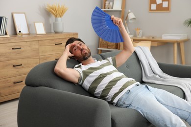 Photo of Bearded man waving blue hand fan to cool himself on sofa at home