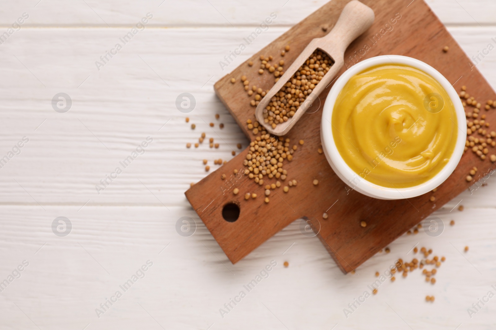 Photo of Bowl with delicious mustard and seeds on white wooden table, flat lay. Space for text