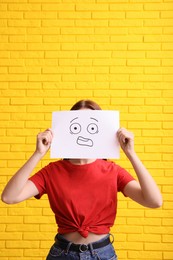 Photo of Woman hiding behind sheet of paper with scared face near yellow brick wall