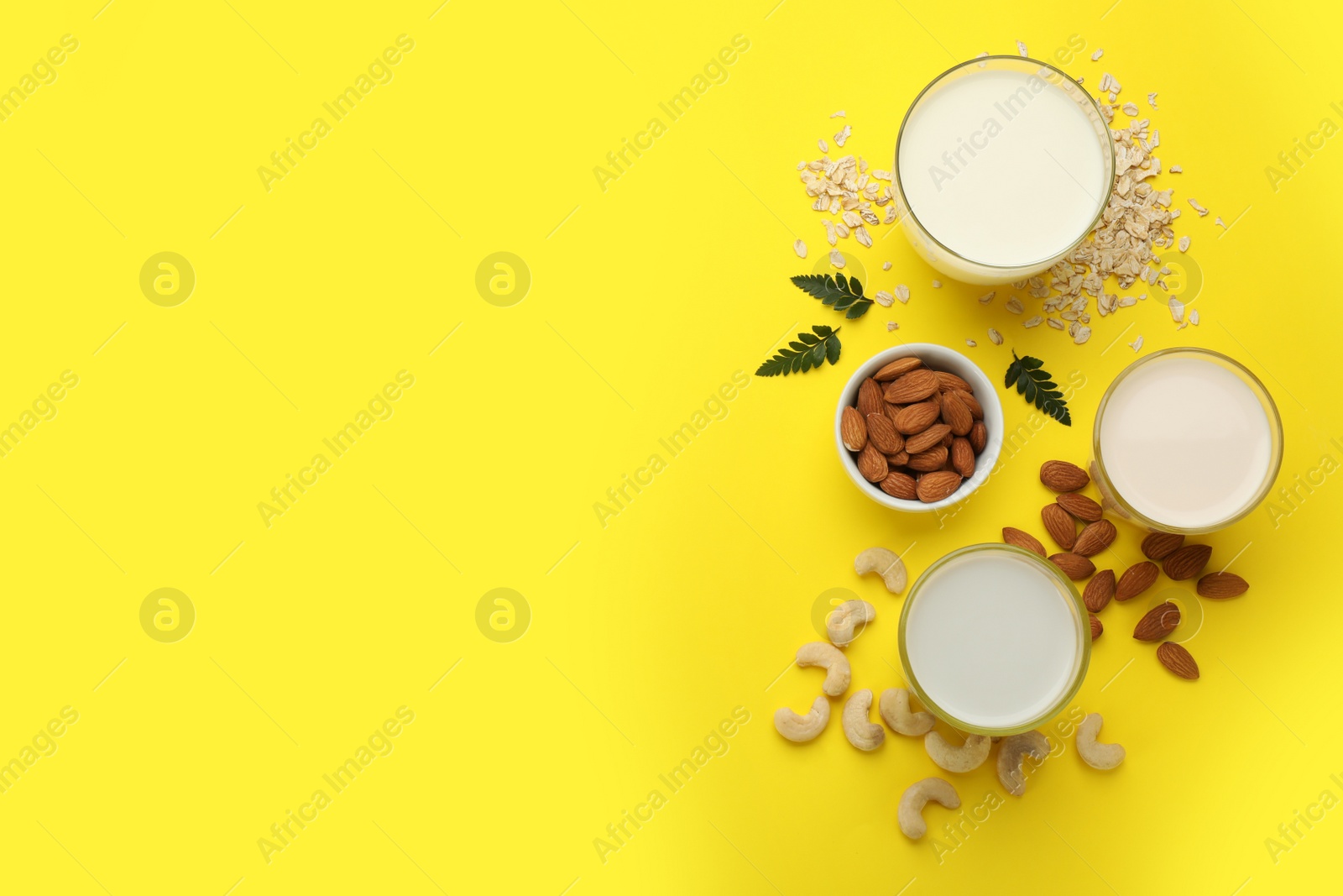 Photo of Different vegan milks, oat flakes and nuts on yellow background, flat lay. Space for text