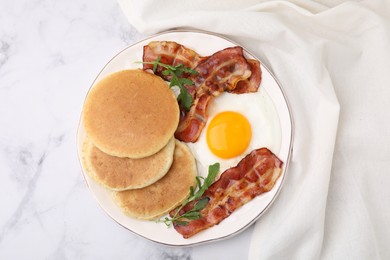 Plate with tasty pancakes, fried egg, arugula and bacon on white marble table, top view