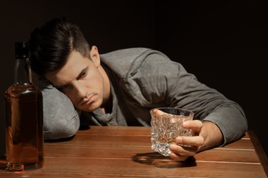 Photo of Addicted man at wooden table against dark background, focus on glass of alcoholic drink