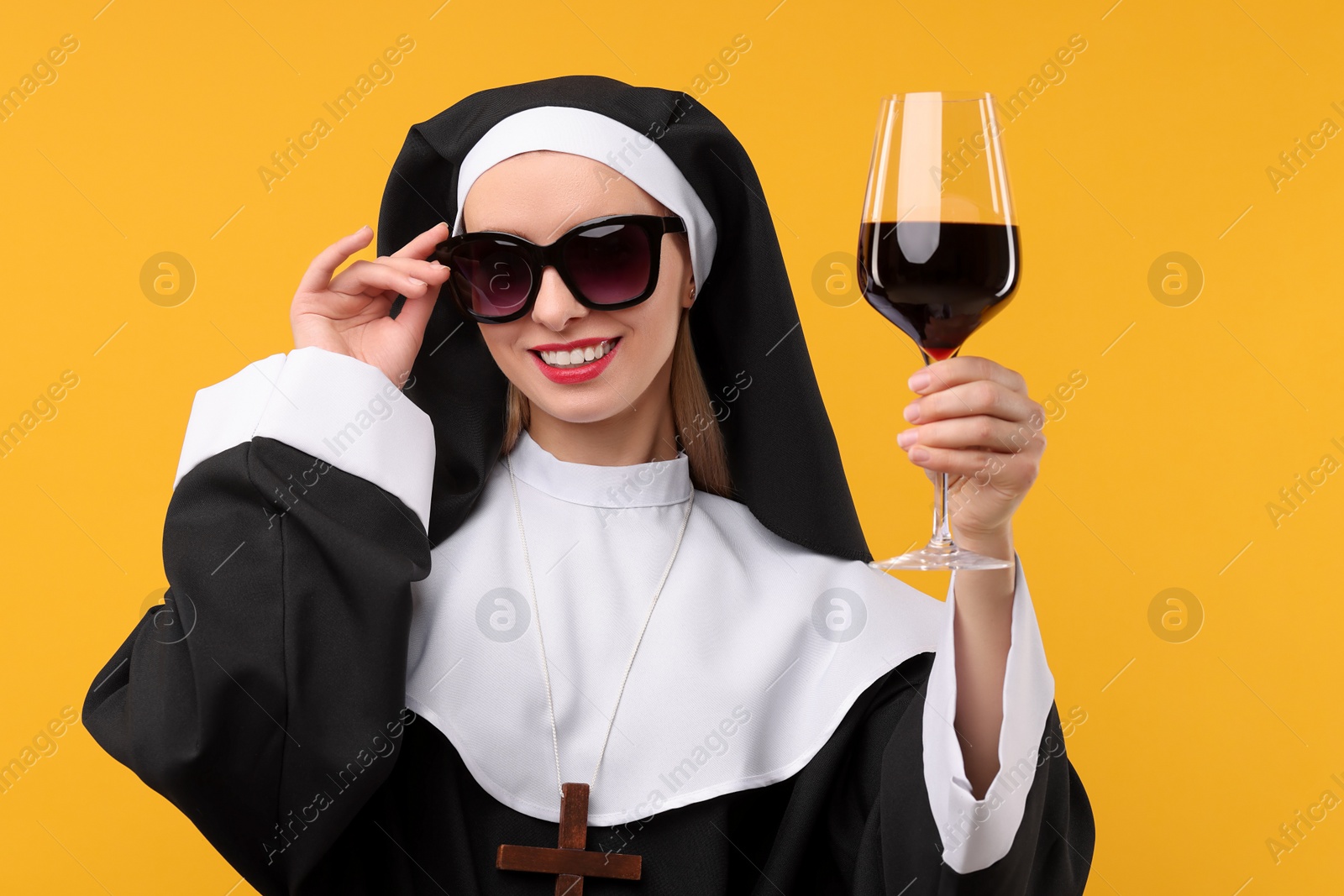 Photo of Happy woman in nun habit and sunglasses holding glass of wine against orange background. Sexy costume