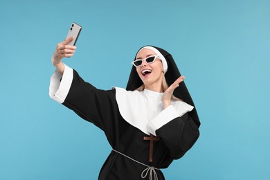 Happy woman in nun habit taking selfie against light blue background
