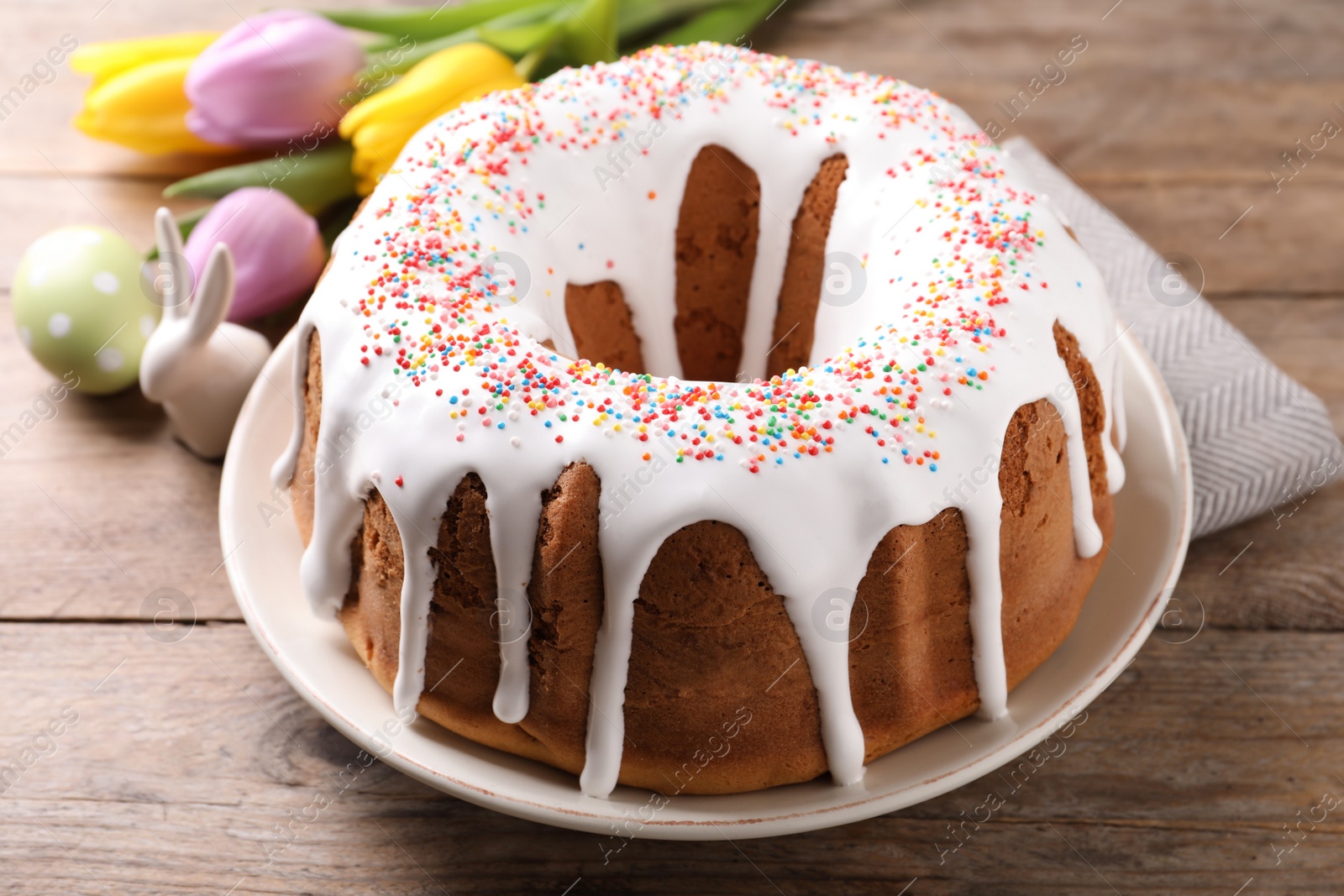 Photo of Glazed Easter cake with sprinkles, decorative bunny, painted egg and tulips on wooden table, closeup