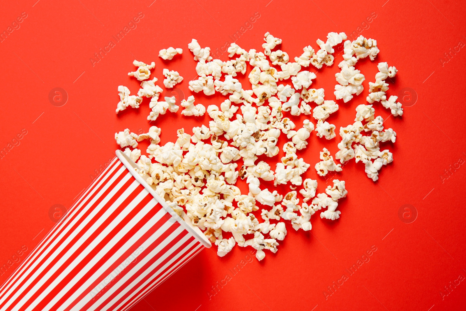 Photo of Overturned paper cup with delicious popcorn on red background, top view