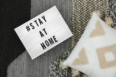 Pillow and lightbox with hashtag STAY AT HOME on warm knitted blanket, flat lay. Message to promote self-isolation during COVID‑19 pandemic