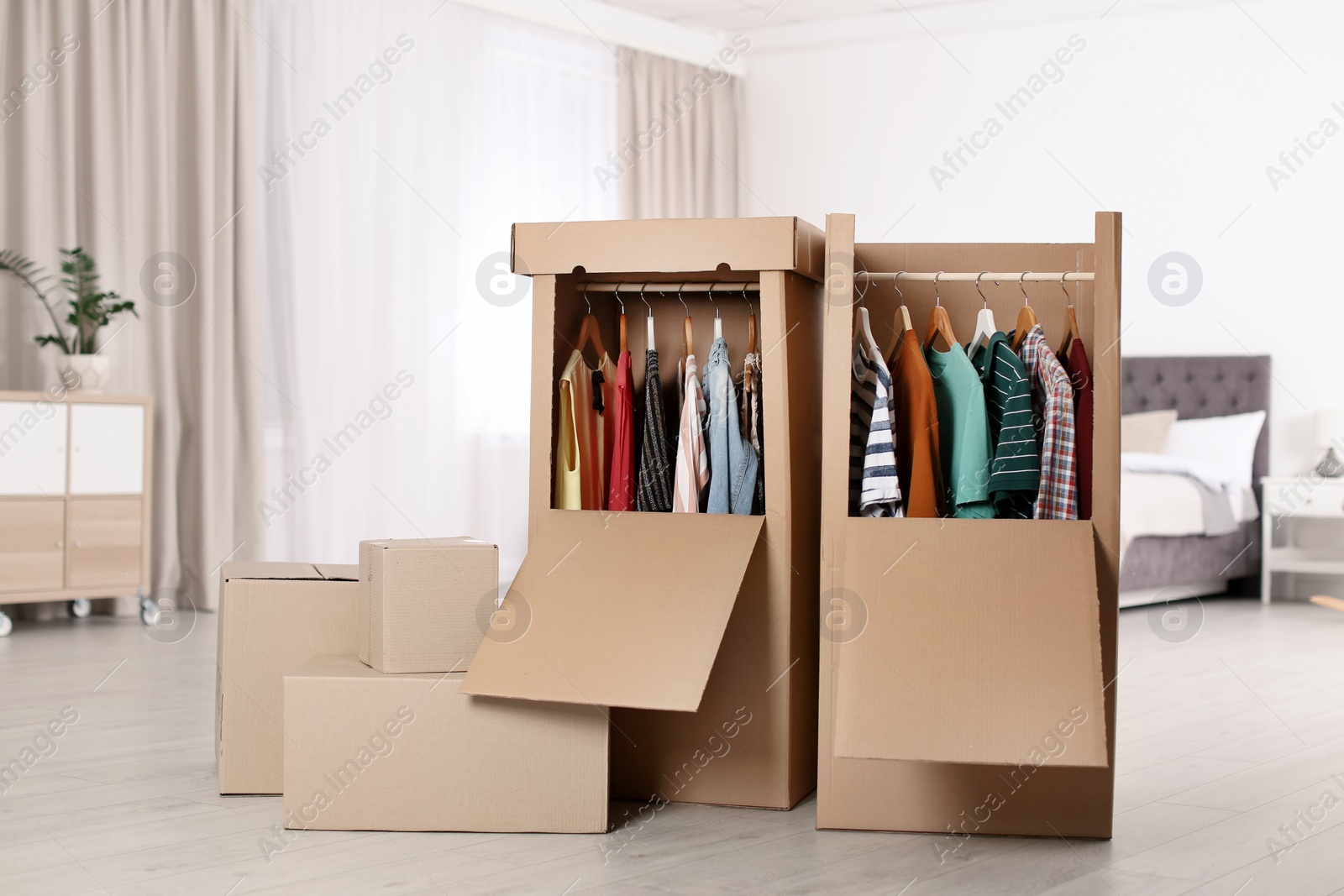 Photo of Cardboard wardrobe boxes with clothes on hangers in bedroom. Space for text