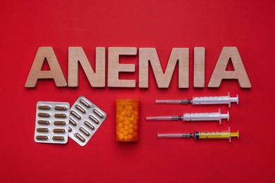 Photo of Word Anemia made with wooden letters, pills and syringes on red background, flat lay