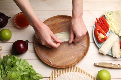 Photo of Making delicious spring rolls. Woman wrapping fresh vegetables into rice paper at wooden table, flat lay