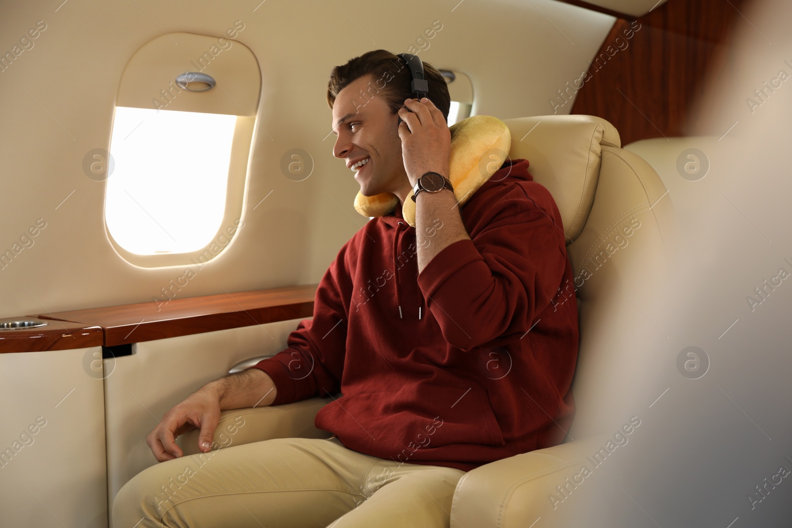 Photo of Young man with travel pillow and headphones listening to music in airplane during flight