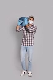 Photo of Courier in medical mask holding bottle for water cooler on light grey background. Delivery during coronavirus quarantine