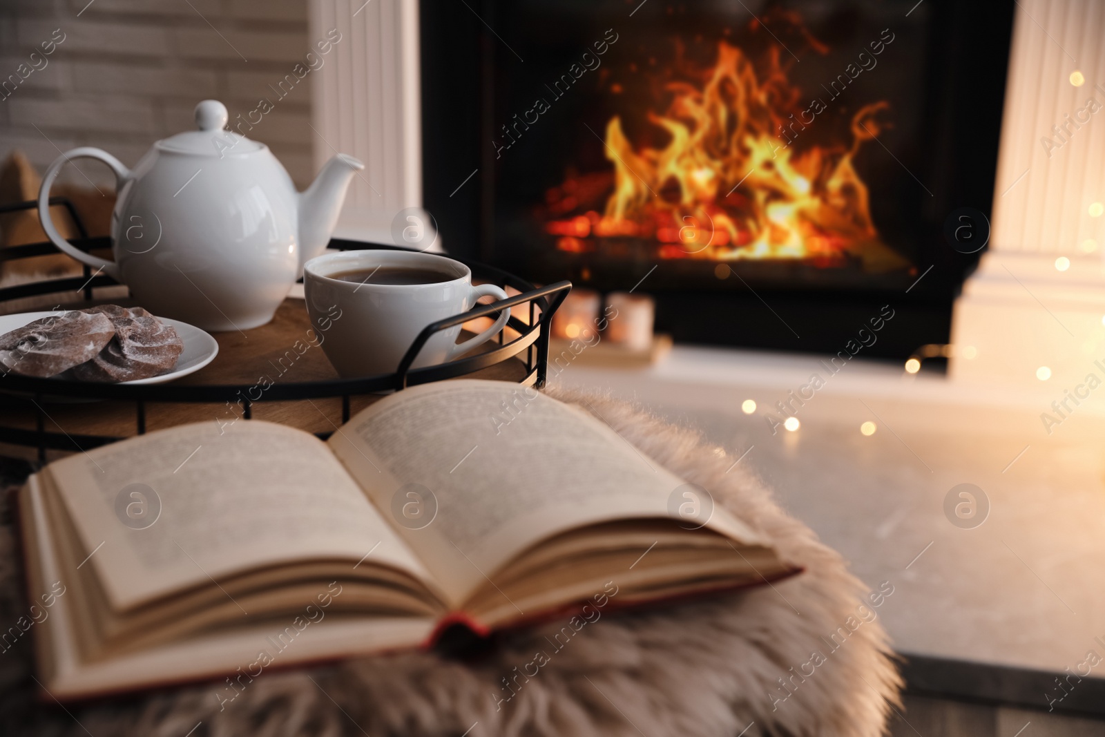 Photo of Cup of tea, cookies and book on faux fur near fireplace indoors, space for text. Cozy atmosphere