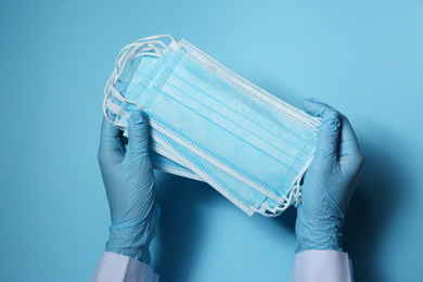 Photo of Doctor in latex gloves holding disposable face masks on light blue background, closeup. Protective measures during coronavirus quarantine