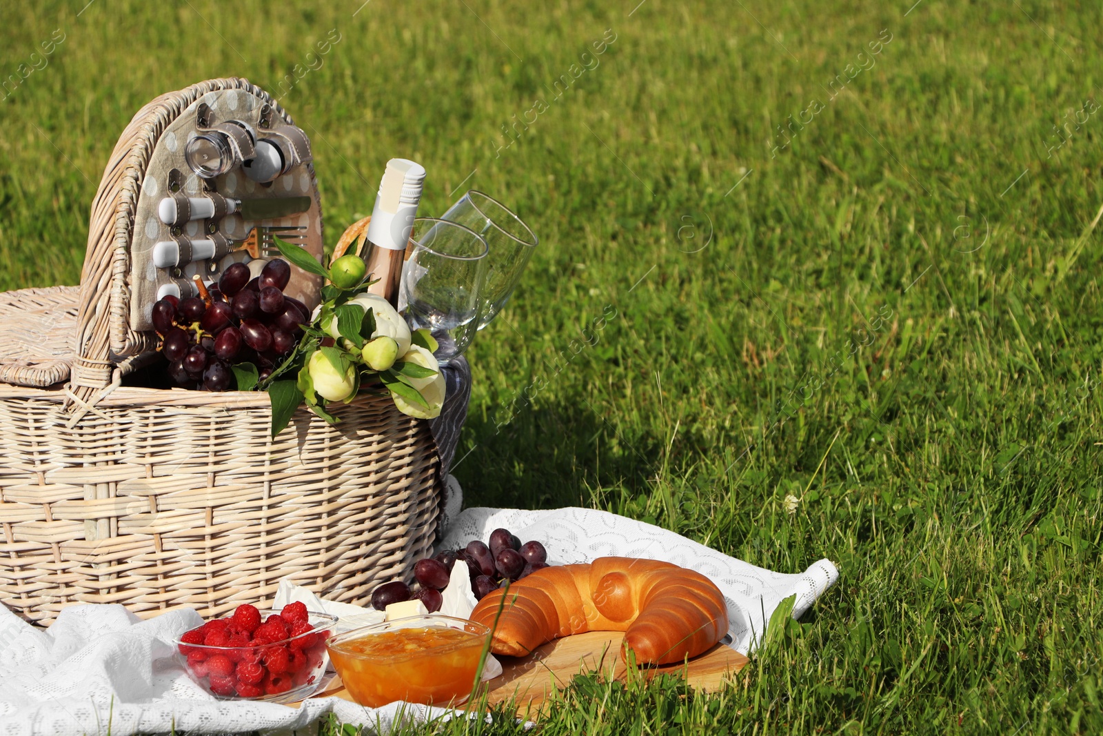 Photo of Picnic blanket with tasty food, flowers, basket and cider on green grass outdoors. Space for text