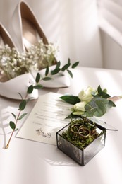 Beautiful wedding rings in glass box, boutonniere, bride's shoes and invitation on white table indoors